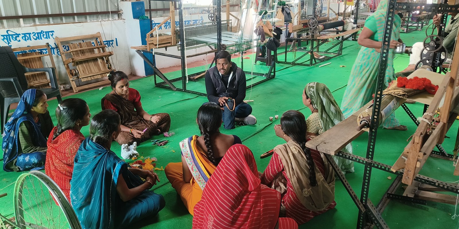 Village women sitting in a circle on the floor of a weaving shed during a training session, looms standing behind them