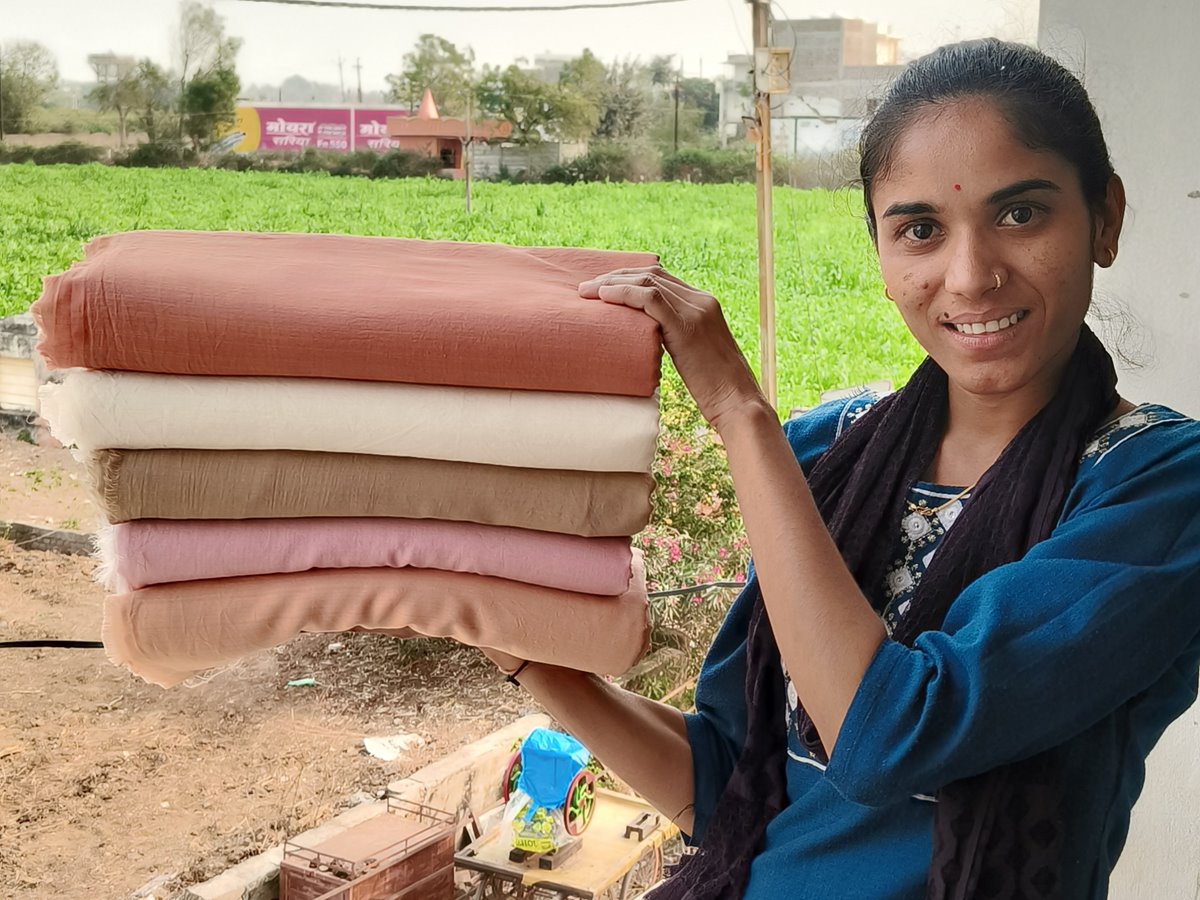 A woman smiling as she holds up a stack of folded handwoven fabric in rust, ivory, tan, pink and peach