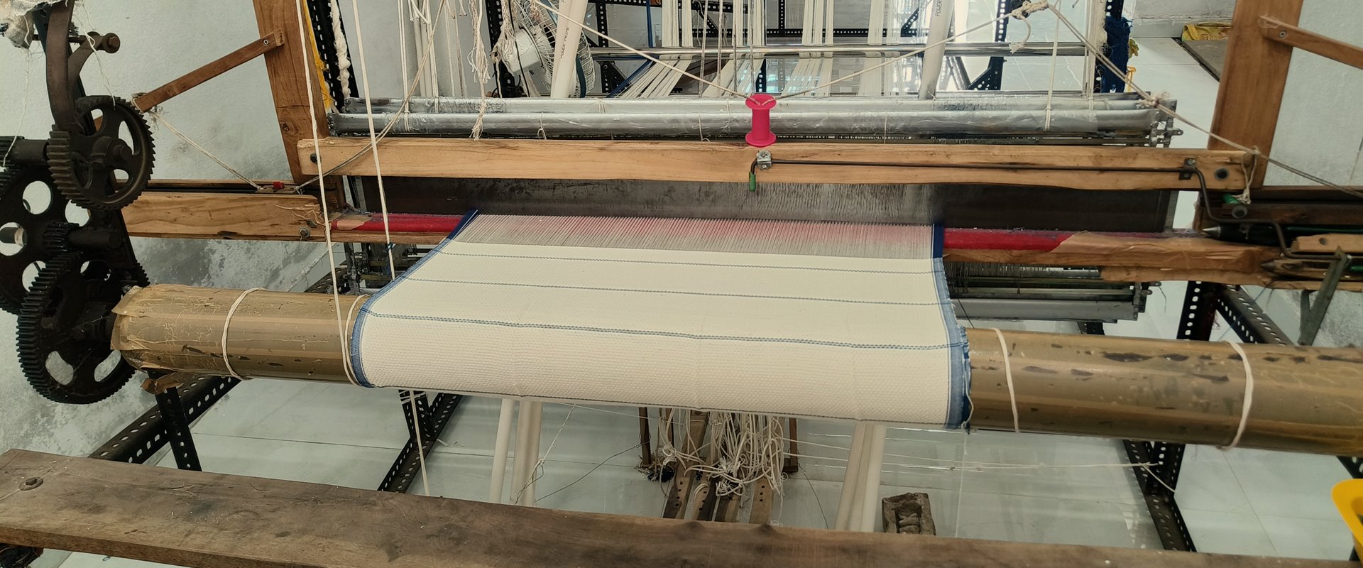 A handloom in our weaving shed, with ivory cloth on the beam and the warp threads rising to the heddles