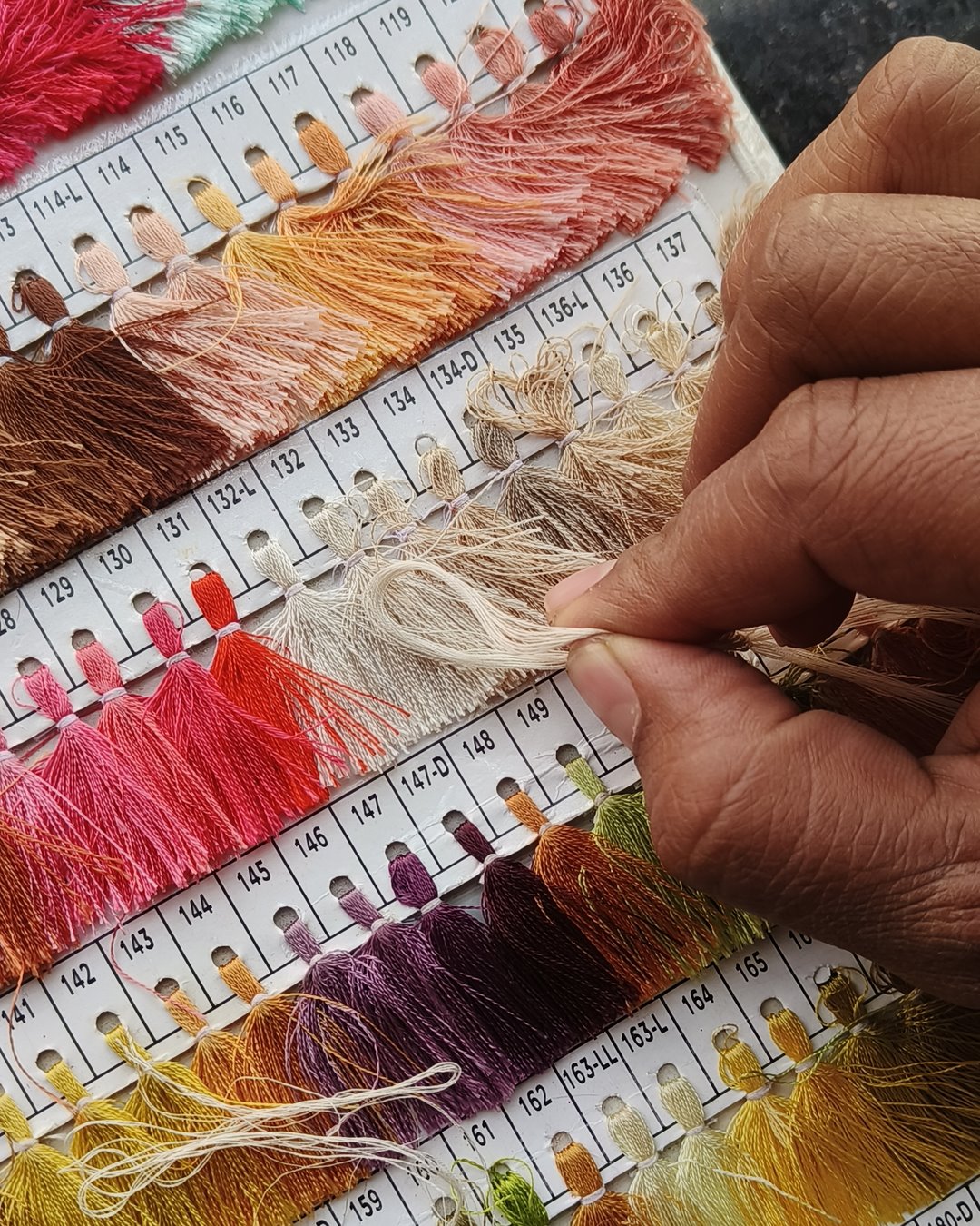 A hand holding a strand of ivory yarn against a card of numbered thread shades