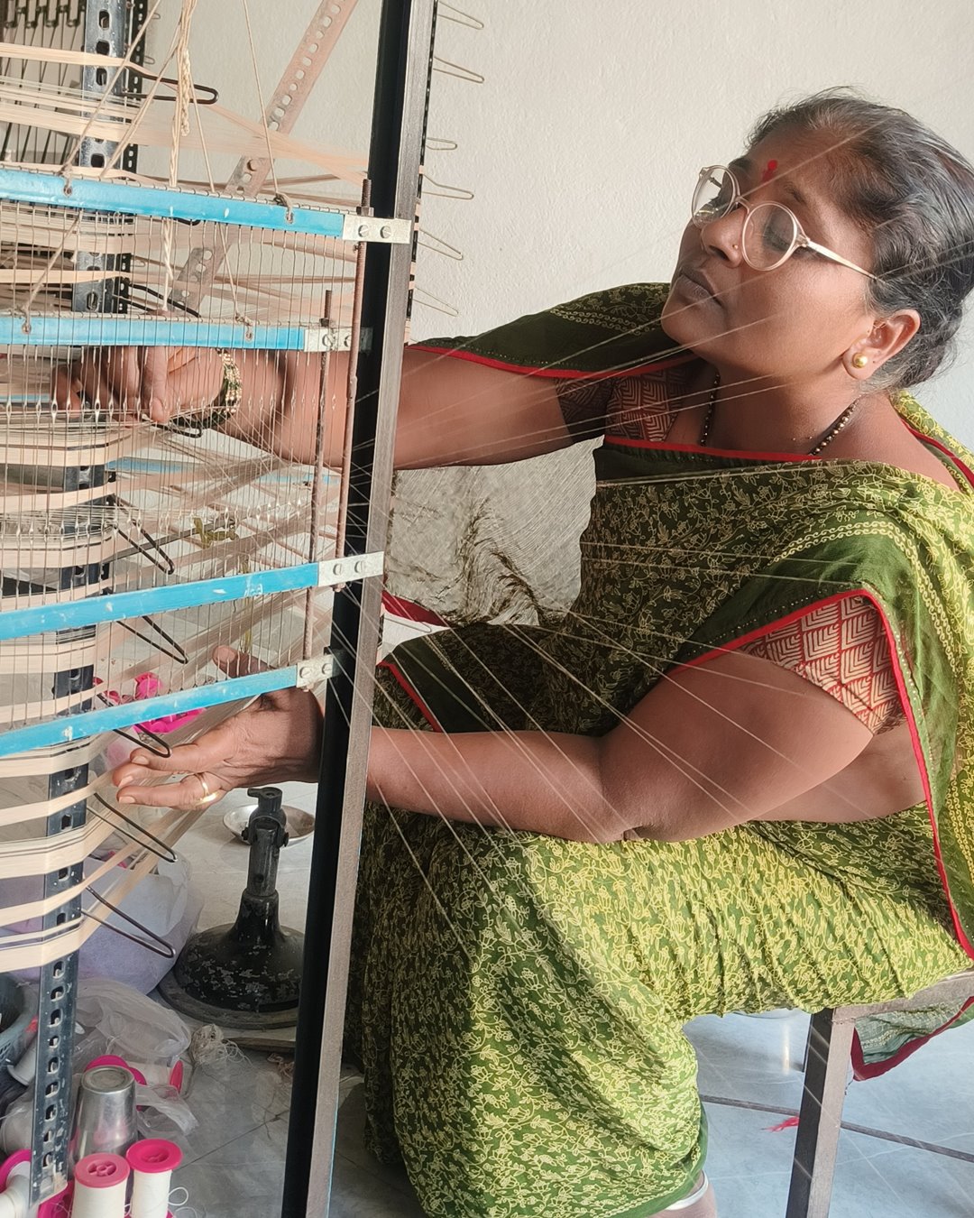 A woman in a green sari guiding yarn through the creel of a warping frame