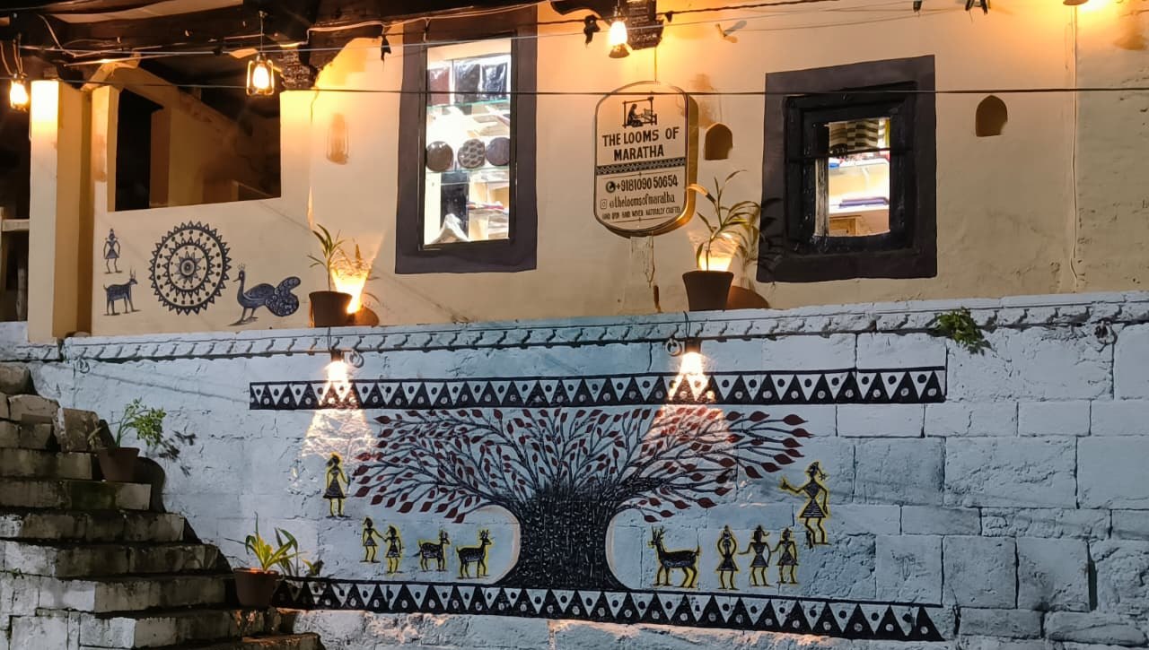 The Looms of Maratha shop in Maheshwar at dusk, its signboard lit above a hand-painted Warli mural